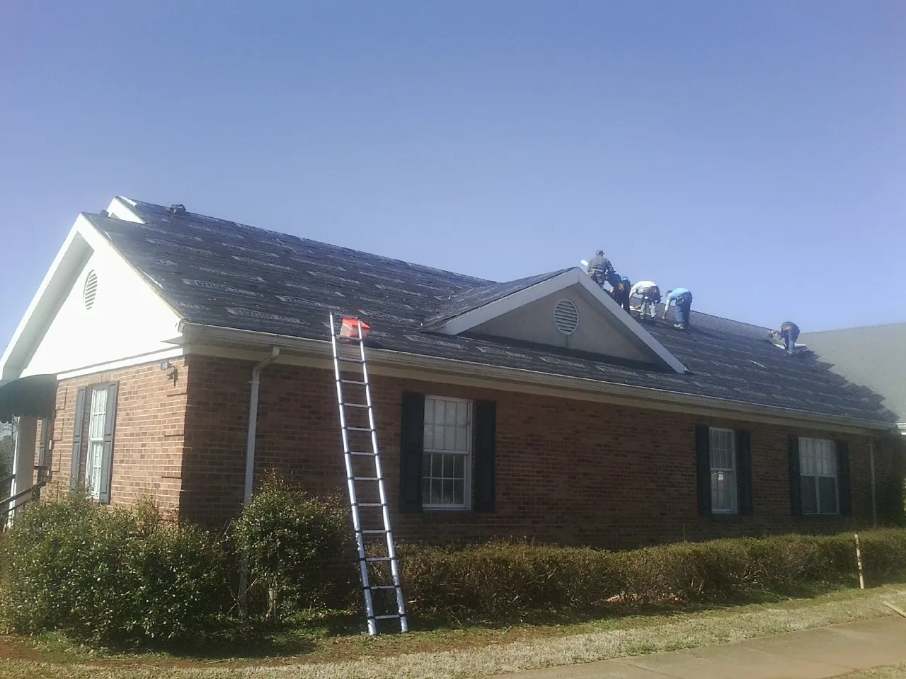 Crew tearing shingles off a brick ranch roof with a ladder set — Metal Roof Repair work by our crew in Severn