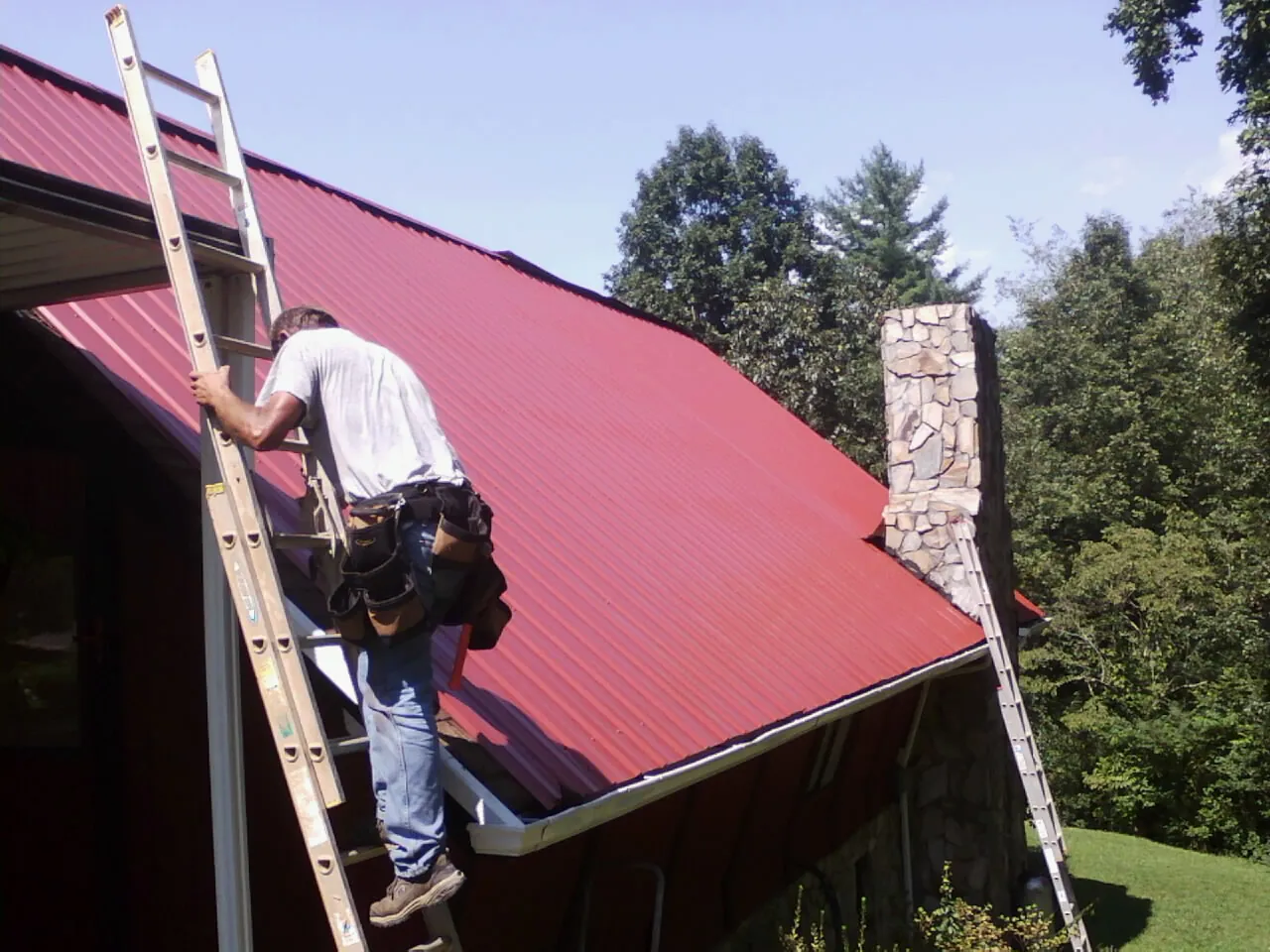 Roofer on a ladder beside a red metal roof and stone chimney — Soffit & Fascia Repair by Roofing Brothers in Severn